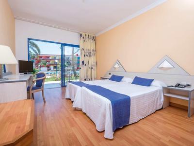 Helles Zimmer mit Doppelbett, blau-weißer Bettdecke, Schreibtisch, Stuhl und Balkon mit Gartenblick.