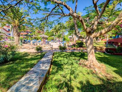 Slunná hotelová zahrada s dlážděnou cestou, velkými stromy a zelenými trávníky.