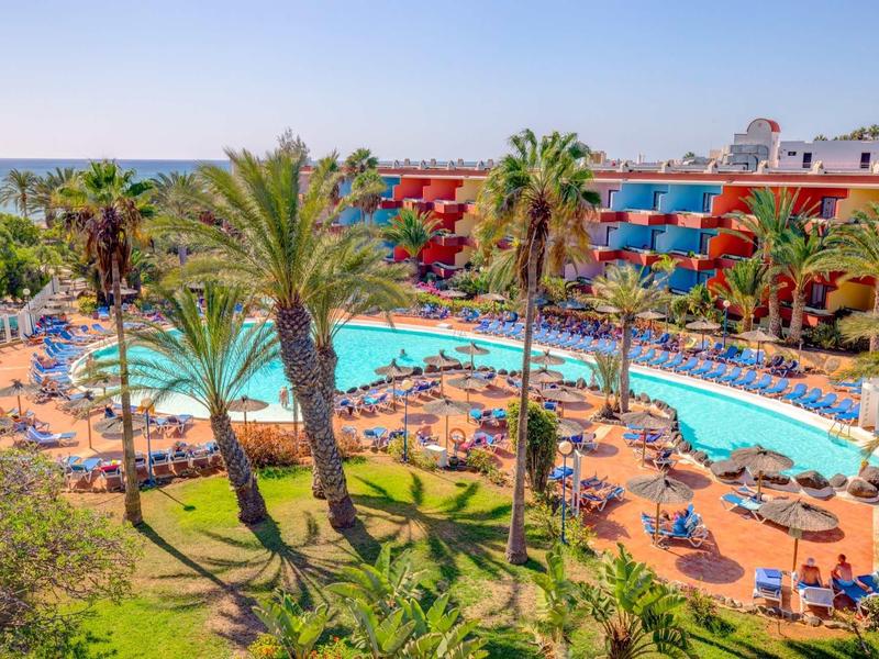 Hotelový bazén s palmami a lehátky, v pozadí moře a vícepatrová budova.