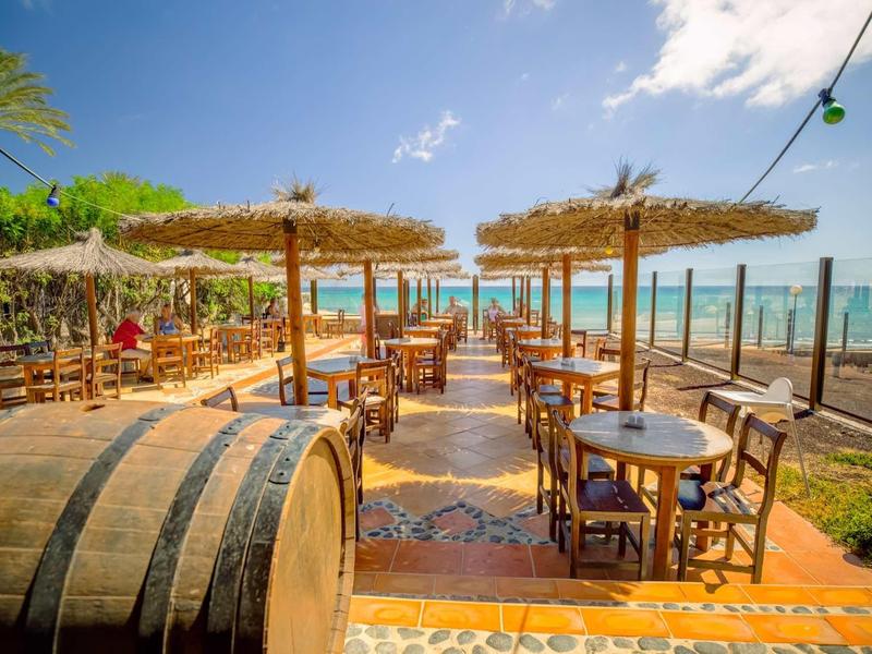 Venkovní restaurace se slámovými slunečníky, dřevěnými stoly a výhledem na moře za jasné oblohy.