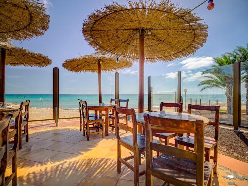 Terasa s dřevěnými stoly a židlemi pod slámovými slunečníky na slunné pláži.