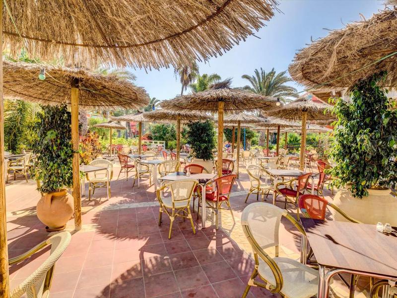 Venkovní restaurace s slaměnými slunečníky, barevnými židlemi a stoly ve slunečném prostředí.