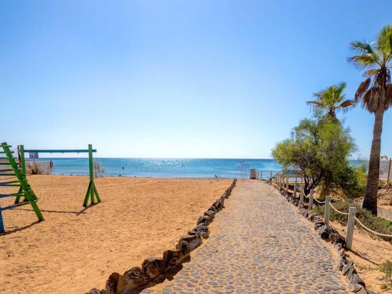 Písčitá dětská hřiště s houpačkami vedle dlážděné cesty vedoucí k moři za jasné oblohy.