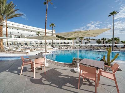 Piscine d'hôtel moderne avec chaises longues, parasols blancs et palmiers sous un ciel bleu.