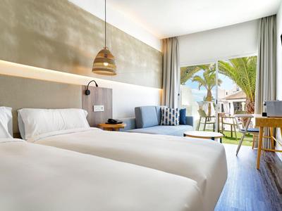 Chambre d'hôtel lumineuse avec deux lits, coin salon et vue sur terrasse et palmiers.