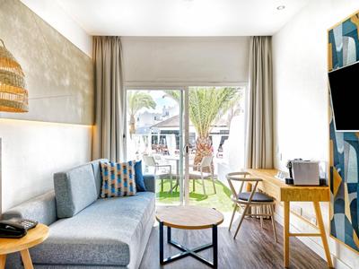 Chambre d'hôtel moderne avec canapé, bureau, TV et vue sur terrasse avec jardin.