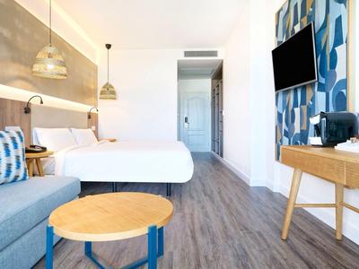Chambre d'hôtel moderne avec lit double, canapé, sol en bois et télévision murale.