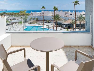 Balcon avec table et chaises donnant sur la piscine, les palmiers et la mer sous un ciel clair.