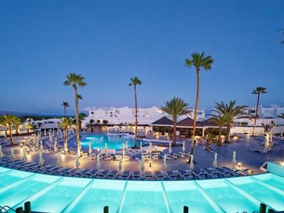 Piscine d'hôtel éclairée avec des palmiers et des bâtiments blancs au coucher du soleil au bord de la mer.