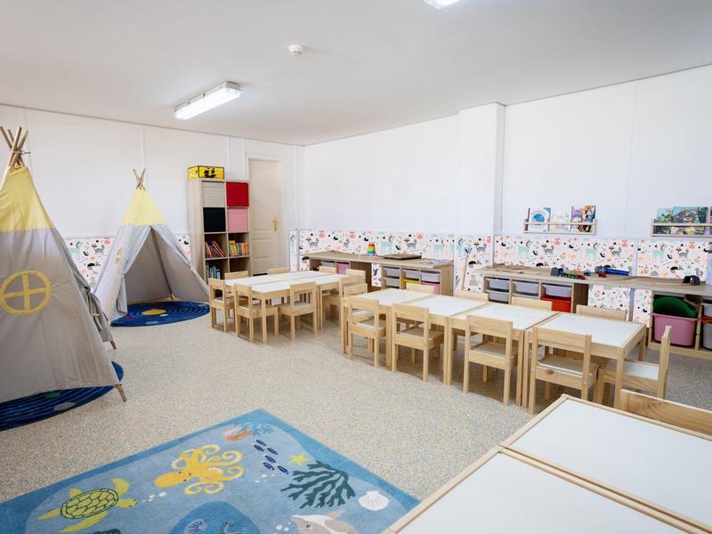 Světla a čistá školková místnost s herními stany, stoly a židlemi a barevným kobercem.