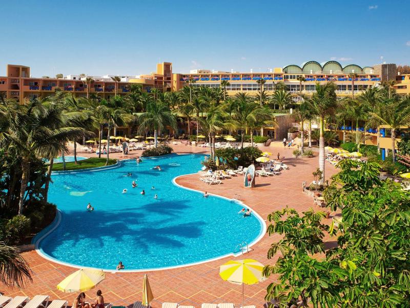 Velký venkovní bazén s palmami a lehátky před rozlehlým hotelem pod modrou oblohou.