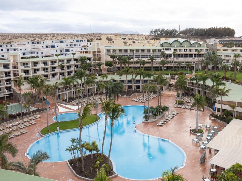 Velký hotel s zakřiveným bazénem obklopený palmami a lehátky.