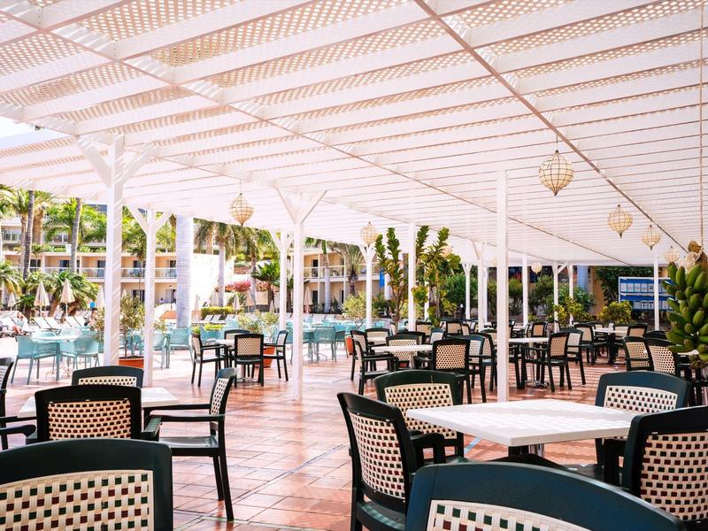 Velká zastřešená venkovní plocha s mnoha stoly a židlemi, obklopená palmami.