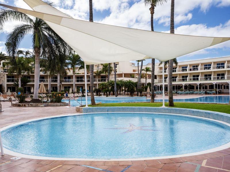 Malý kulatý bazén se sluneční plachtou, obklopený lehátky a palmami v hotelu.