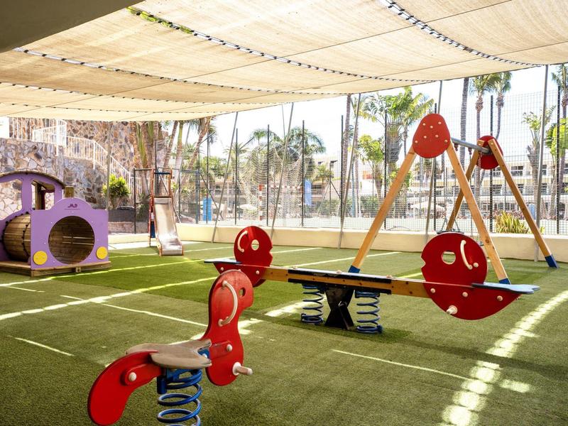 Zastřežené hřiště s barevnými houpačkami, houpačkou a horolezeckou konstrukcí na umělém trávníku.