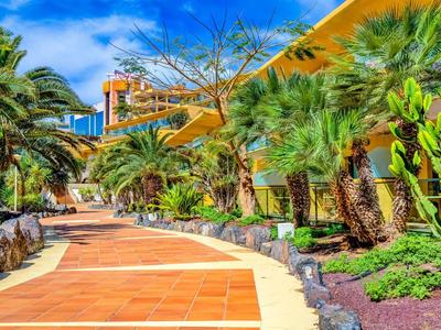 Weg mit roten Fliesen, umgeben von Palmen und tropischer Vegetation unter blauem Himmel.