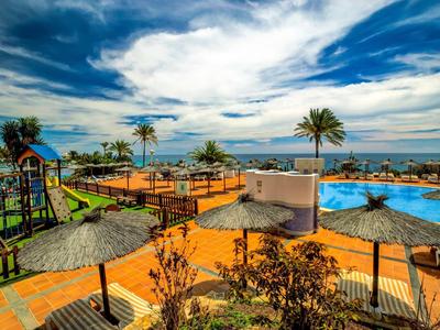 Hotelová bazénová oblast s slunečníky, palmami a výhledem na moře za oblačného nebe.