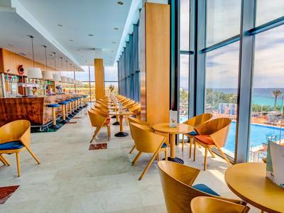 Moderní kavárna s dřevěným nábytkem a panoramatickými okny s výhledem na bazén a moře.