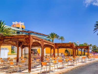 Sonnige Terrasse mit Holzpergola, Tischen und Stühlen neben einem gelben Gebäude und Palmen.