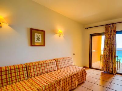 Světlý hotelový pokoj s kostkovanou pohovkou, nástěnnými lampami a výhledem na moře skrz dveře.