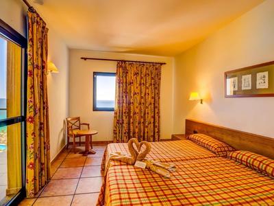 Světle zařízený hotelový pokoj s manželskou postelí, balkonem s výhledem na moře a dekorativními závěsy.