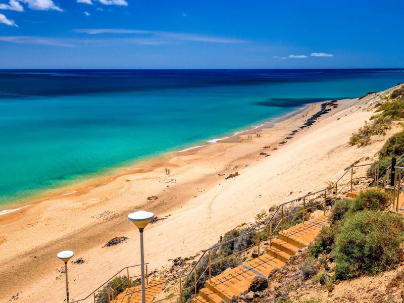Rozlehlá písečná pláž s tyrkysovým mořem a schodištěm mezi dunami.