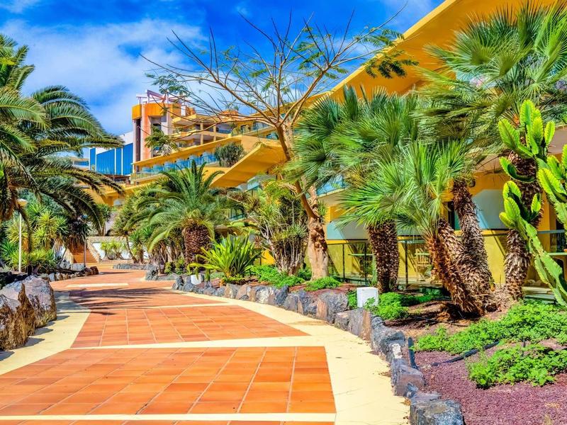 Široká chodba s palmami a tropickou vegetací vedle hotelové budovy.