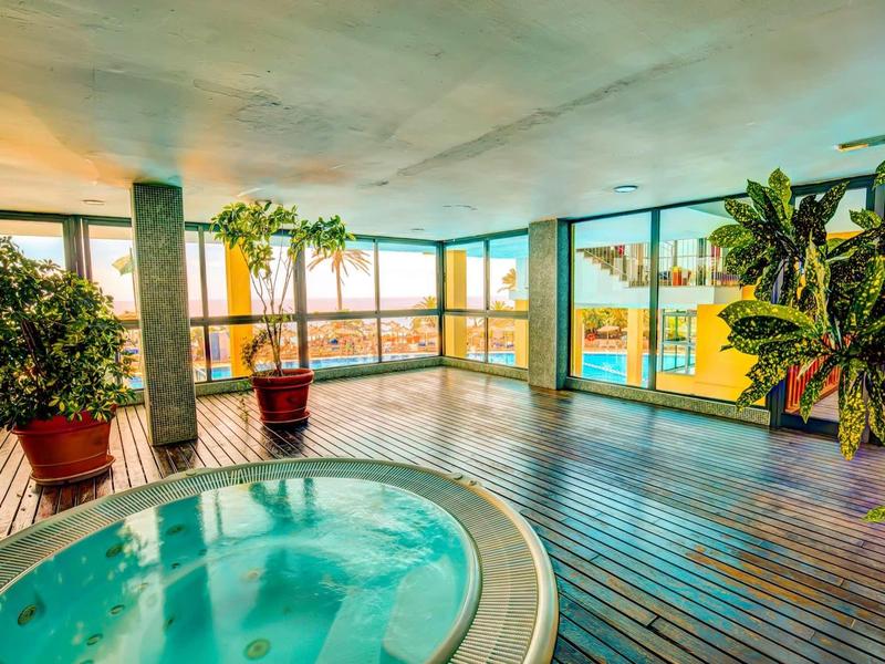 Krytá oblast s vířivkou s dřevěnou podlahou a velkými panoramatickými okny s výhledem na palmy.