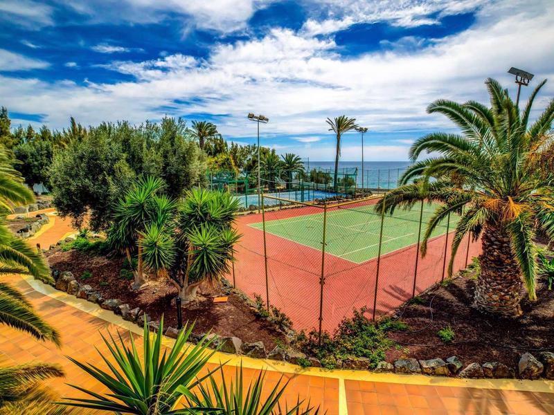 Venkovní tenisový kurt obklopený palmami a tropickou vegetací s výhledem na blízké moře.