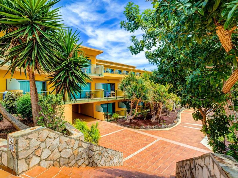 Moderní hotelový komplex se žlutou fasádou, obklopený palmami a modrou oblohou.