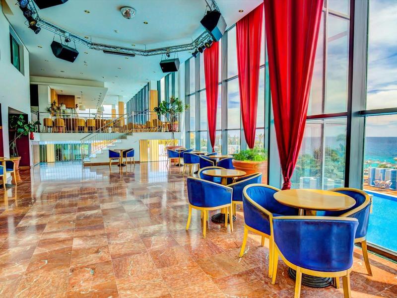Světla hotelová jídelna s velkými okny, červenými závěsy, modrými židlemi a výhledem na moře.