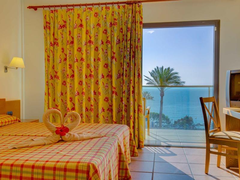 Hotelový pokoj se dvěma postelemi, barevnými závěsy a balkonem s výhledem na moře a palmy.
