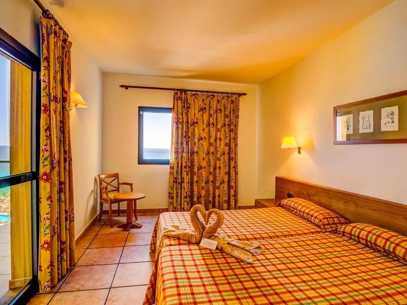 Světle zařízený hotelový pokoj s manželskou postelí, balkonem s výhledem na moře a dekorativními závěsy.