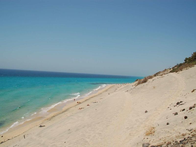 Pláž s jemným pískem a průzračnou tyrkysovou vodou pod modrou oblohou.