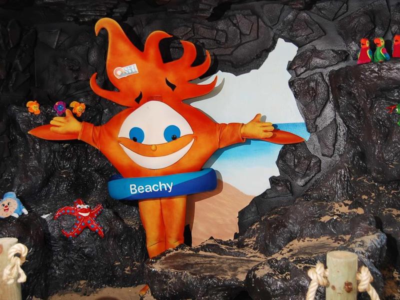 Barevná hotelová maskotka s nápisem 'Beachy' v jeskyni na pláži.