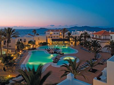 Wieczorny widok na oświetlony hotel z basenem, palmami i widokiem na morze i góry.