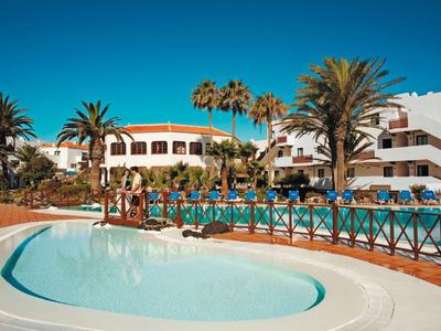 Complexe hôtelier avec plusieurs bâtiments blancs, des palmiers et deux piscines sous un ciel bleu clair.