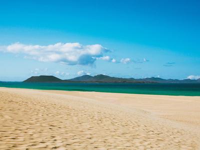 Pusta piaszczysta plaża z niebieskim niebem i zielonymi wyspami na horyzoncie.