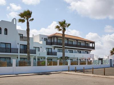 Nowoczesny budynek hotelu z balkonami, palmami i białym ogrodzeniem pod niebieskim niebem.