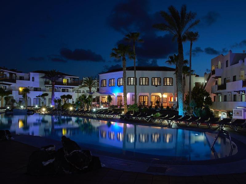 Nocne zdjęcie oświetlonego hotelu z basenem i palmami pod ciemnym niebem.