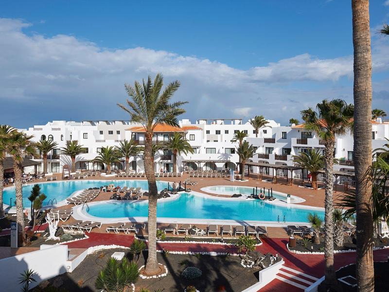 Hotel z białym budynkiem otaczającym duży basen i palmami pod czystym niebem.