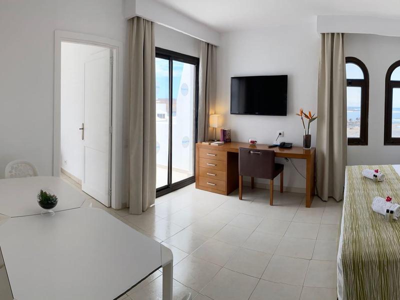 Jasny pokój hotelowy z podwójnym łóżkiem, biurkiem, telewizorem i balkonem z widokiem na zewnątrz.