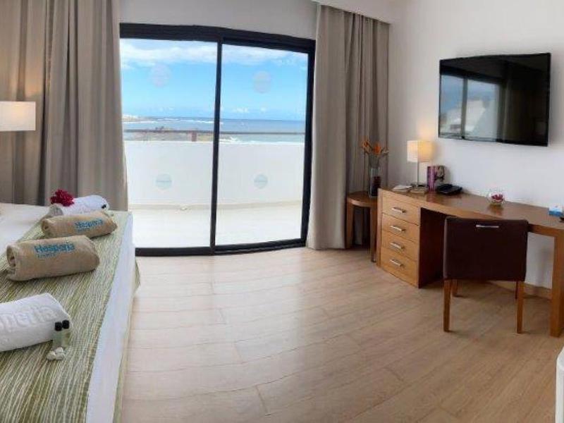 Jasny pokój hotelowy z podwójnym łóżkiem, telewizorem i balkonem z widokiem na morze.