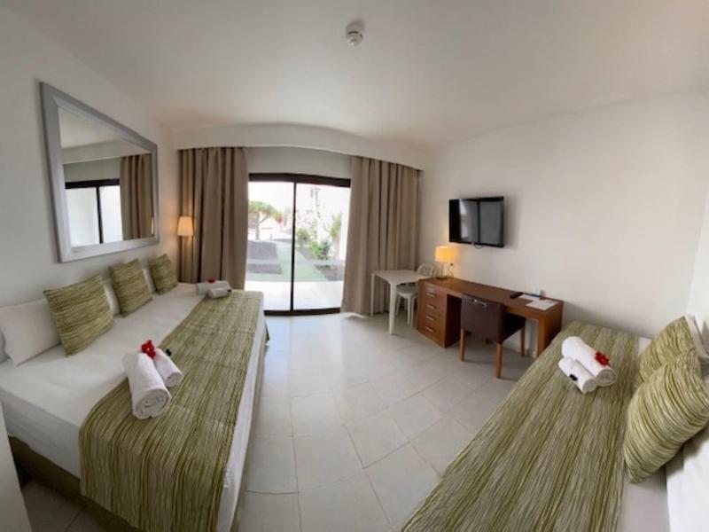 Jasny pokój hotelowy z dwoma łóżkami, biurkiem, telewizorem i dostępem do balkonu.