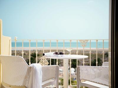 Balkon mit zwei weißen Stühlen und kleinem Tisch, Blick auf Strand und Meer bei klarem Himmel.