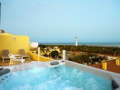 Whirlpool auf Terrasse mit Liegestühlen, gelber Wand und Blick auf Meer und Leuchtturm.