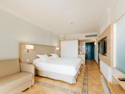 Hell eingerichtetes Hotelzimmer mit Doppelbett, Couch, Flachbildschirm und heller Bodenfliese.