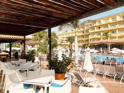 Außenbereich mit Sitzplatz, Holzdach, Pflanzen und Pool mit Liegestühlen vor einem mehrstöckigen Hotel.