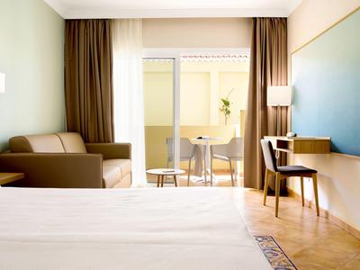 Helles Hotelzimmer mit großem Bett, Sofa, Tisch, Stuhl und Balkon mit Sicht ins Freie.