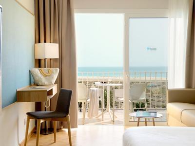 Helles Zimmer mit Stuhl, Schreibtisch, Sofa und Blick auf Balkon und Meer durch große Fenster.